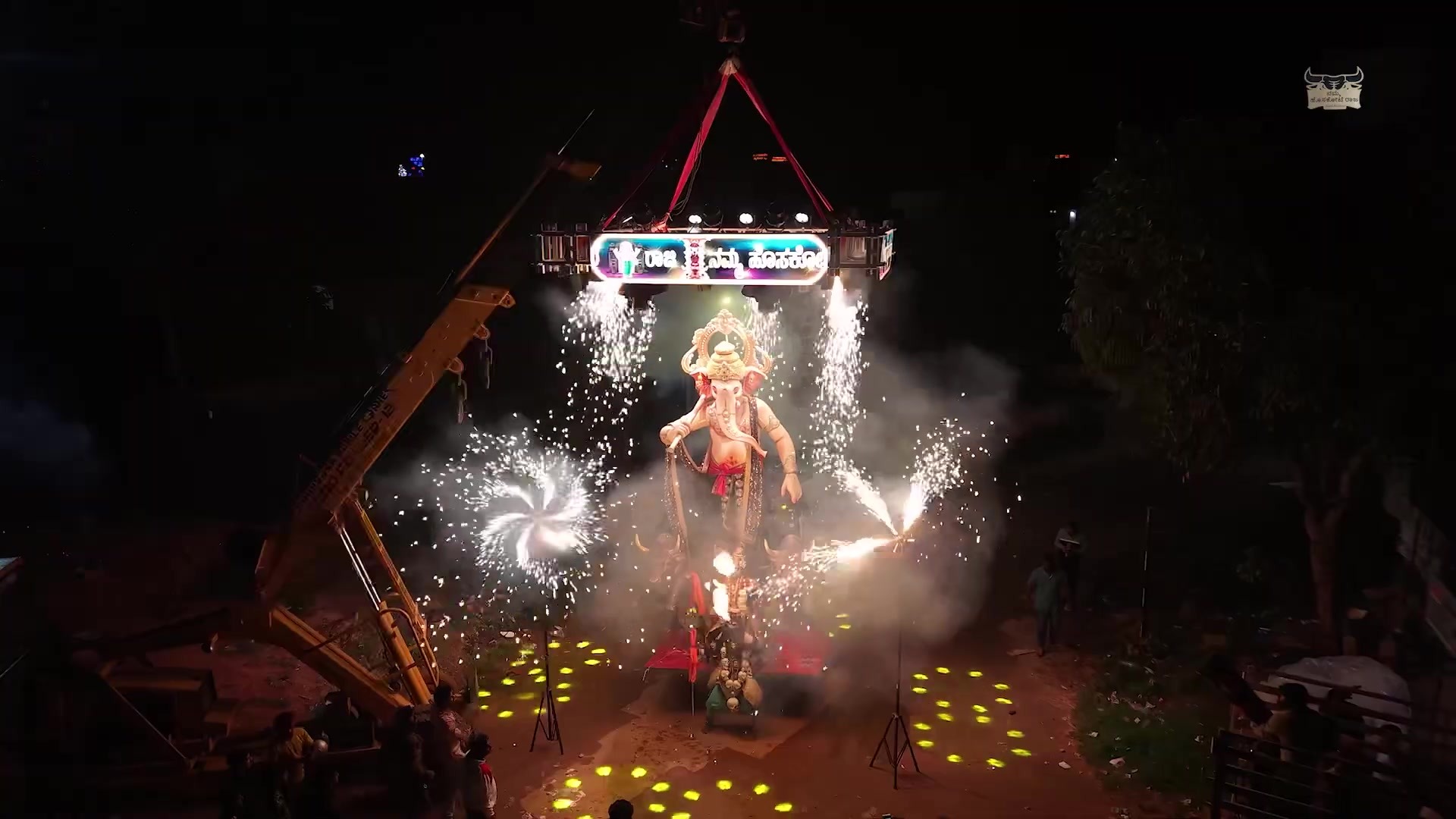 Ganesha idol lifted by crane under a lit gantry, framed by spiral fireworks