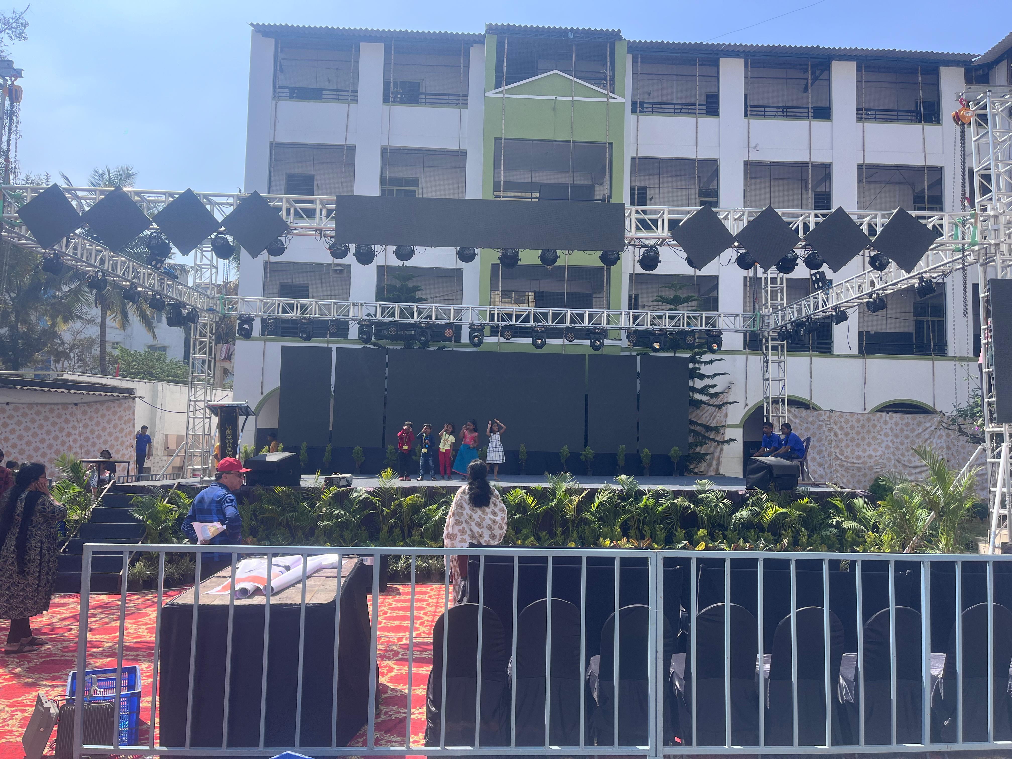 School ground stage with truss-mounted LED panels and moving-head lights, children performing